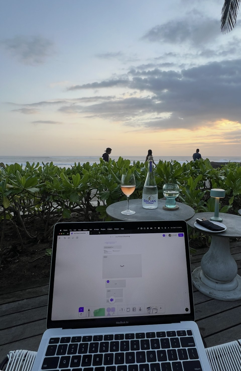 A laptop open to a Figma file, “Jury Flows & Roadmap”, on a beachside deck at sunset, with a glass of rosé and a bottle of water on the table behind it